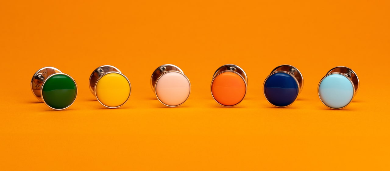 Six pins in green, yellow, pink, orange, dark blue and light blue on an orange studio background
